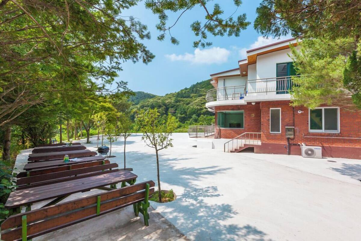 A building with a balcony, a paved area with benches, and a view of a green hillside.