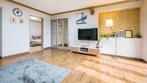 Una sala de estar con piso de madera, un tapete gris, un televisor sobre un mueble blanco, una lámpara y una planta.