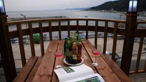 Une table en bois avec vue sur la mer, une bouteille d’eau, une bouteille de jus de fruits, un verre de vin rouge et une assiette de fruits.