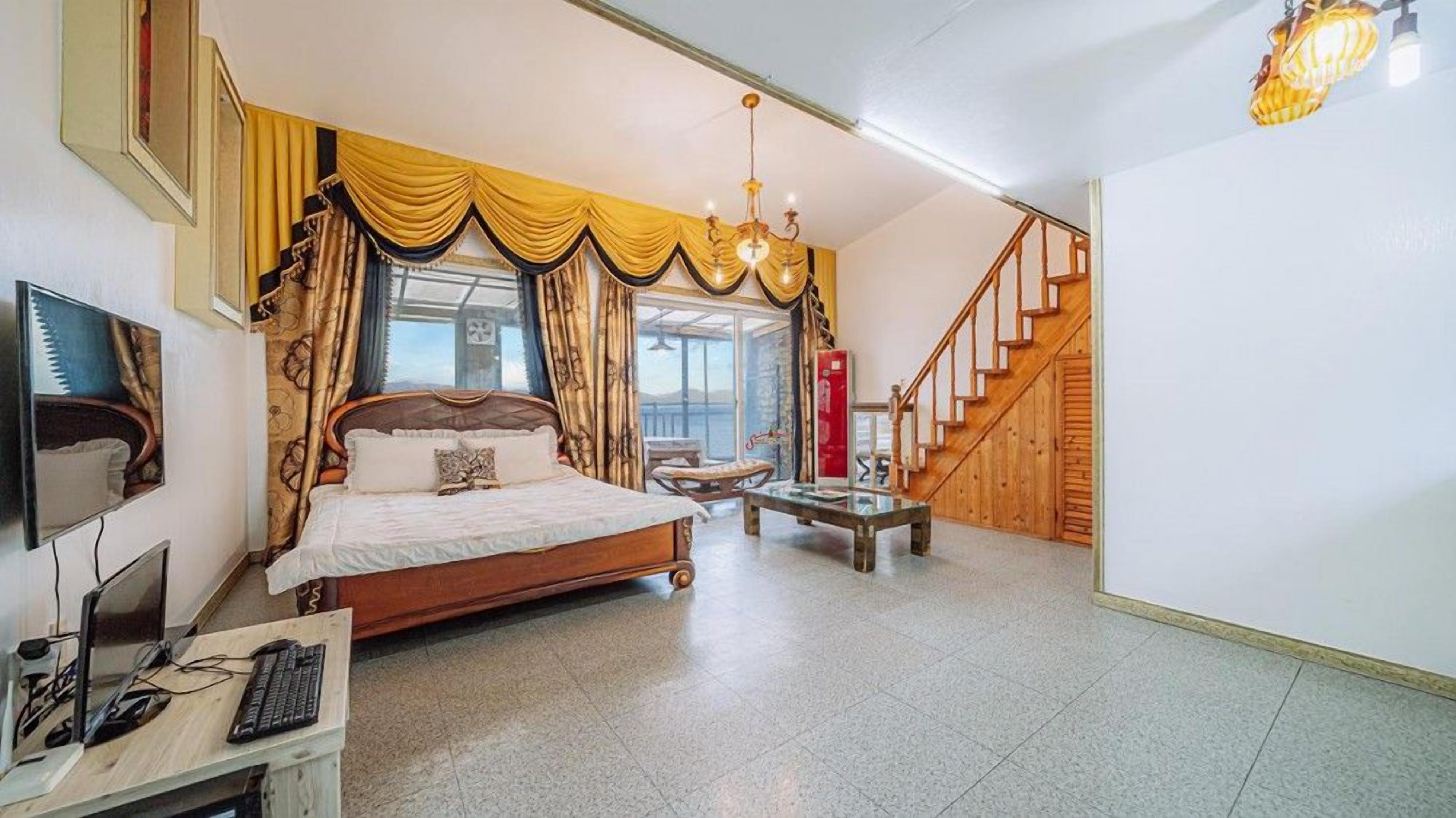 A bedroom with a bed, a desk with a computer, a chair, a table, a staircase, and a window with curtains.