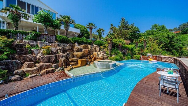 A swimming pool with a wooden deck, a rock waterfall feature, and a lounge area with a table and chairs. - Santamonica Pension (Geoje)