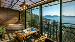 A room with a large glass window, a wooden table, a bathtub, and a hanging light fixture. - Santamonica Pension (Geoje)