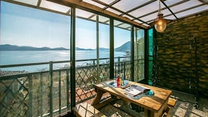 A room with a wooden table and benches, large windows with a view of a body of water and mountains, and a hanging light fixture. - Santamonica Pension (Geoje)
