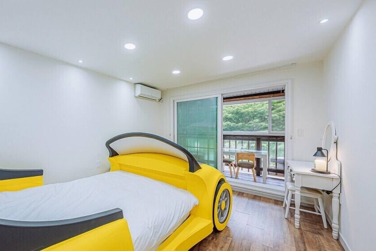 A bedroom with a bed featuring a car-like design, a desk with a chair, and a large window with a view of greenery.
