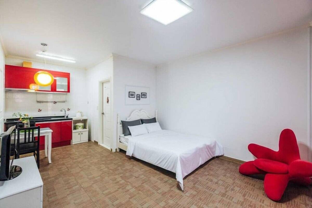 A compact room with a bed, a desk with a chair, a red chair, and a kitchenette.
