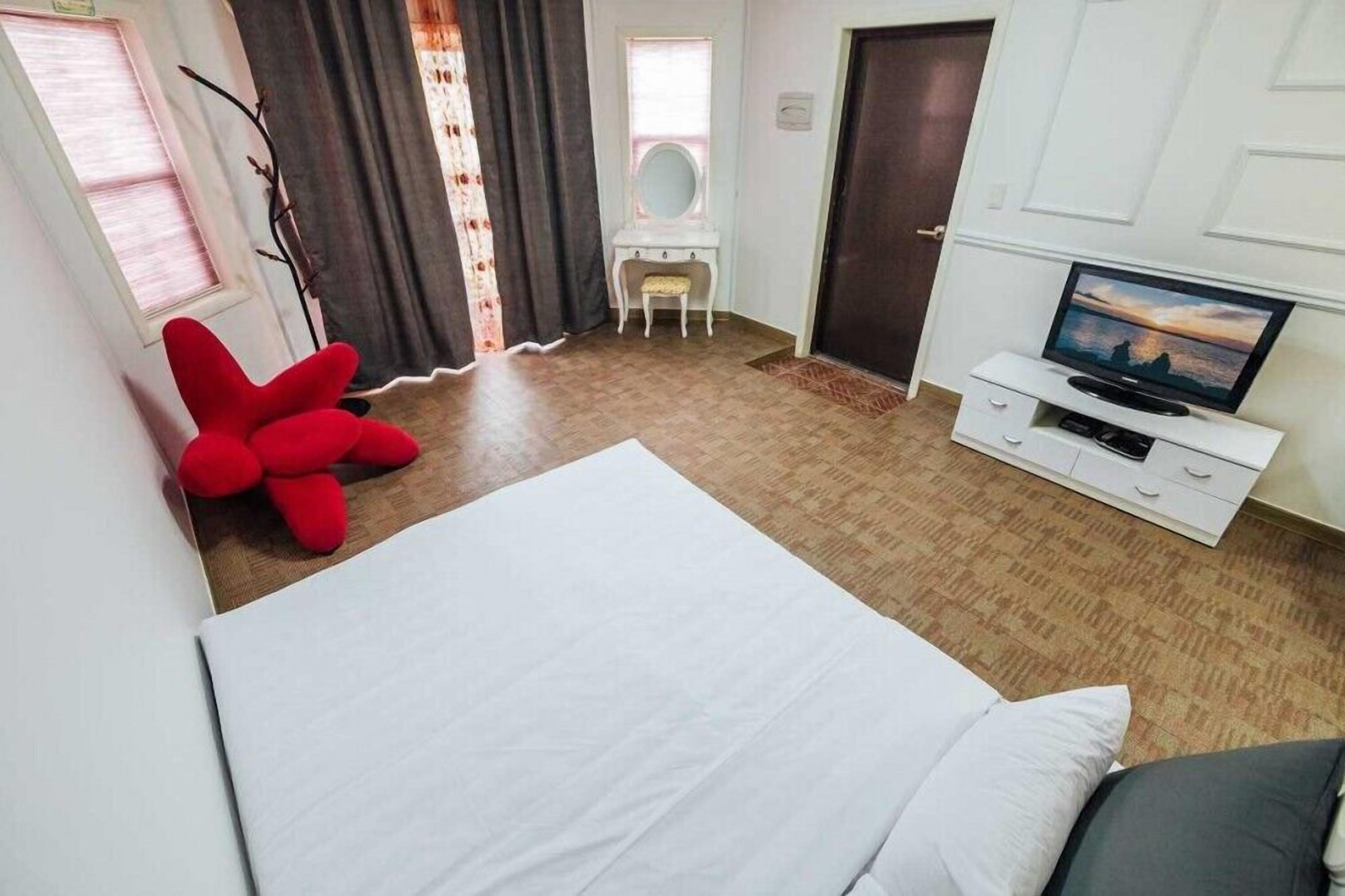 A bedroom with a bed, a TV on a white cabinet, a red chair, and a small desk with a mirror.