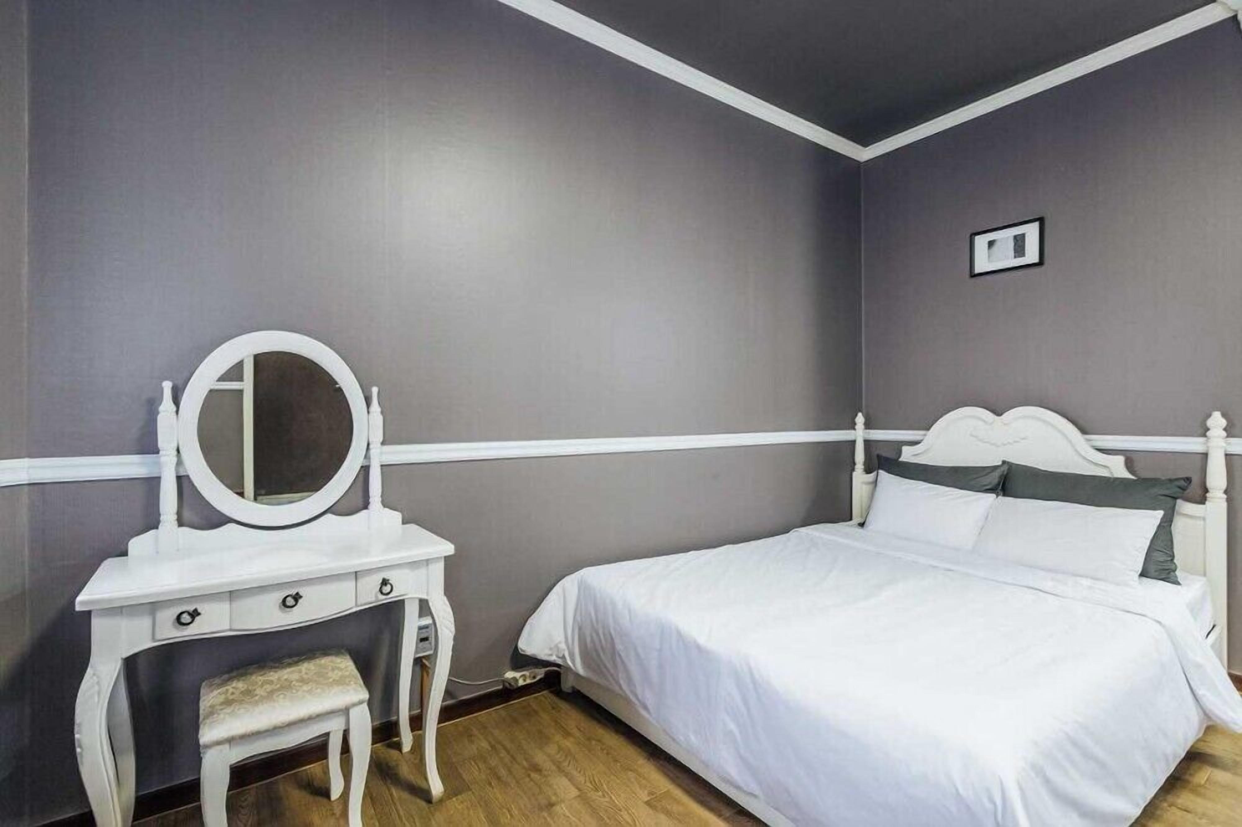 A bedroom with a white vanity, a round mirror, a small bench, a bed with white bedding, and a grey wall.