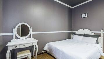 A bedroom with a white vanity, a round mirror, a small bench, a bed with white bedding, and a grey wall.