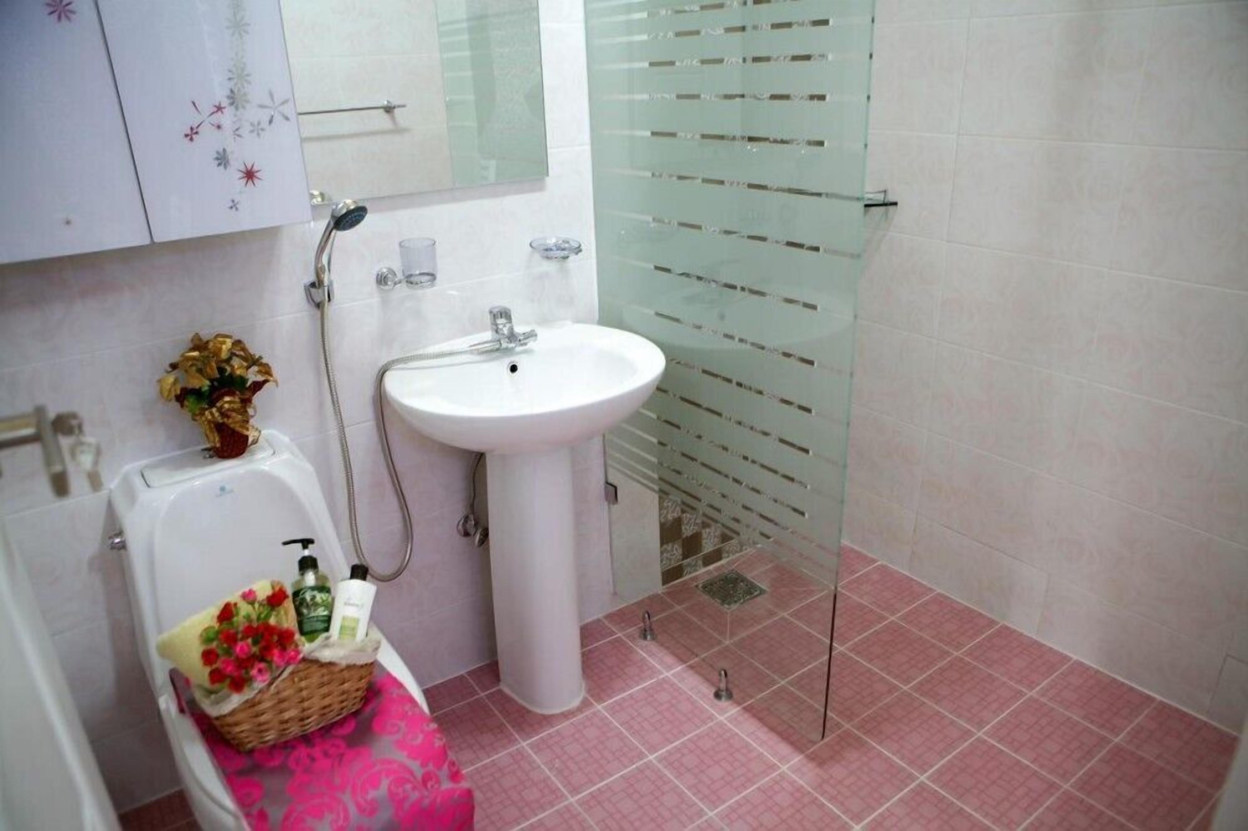 Un baño con un lavamanos blanco, cabina de ducha con mampara de vidrio y un inodoro con un cesto de flores.