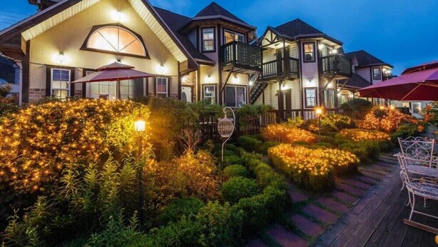 A large, multi-story house with a well-lit exterior, surrounded by landscaped gardens and outdoor seating areas. - Indigo Pension (Hwaseong)
