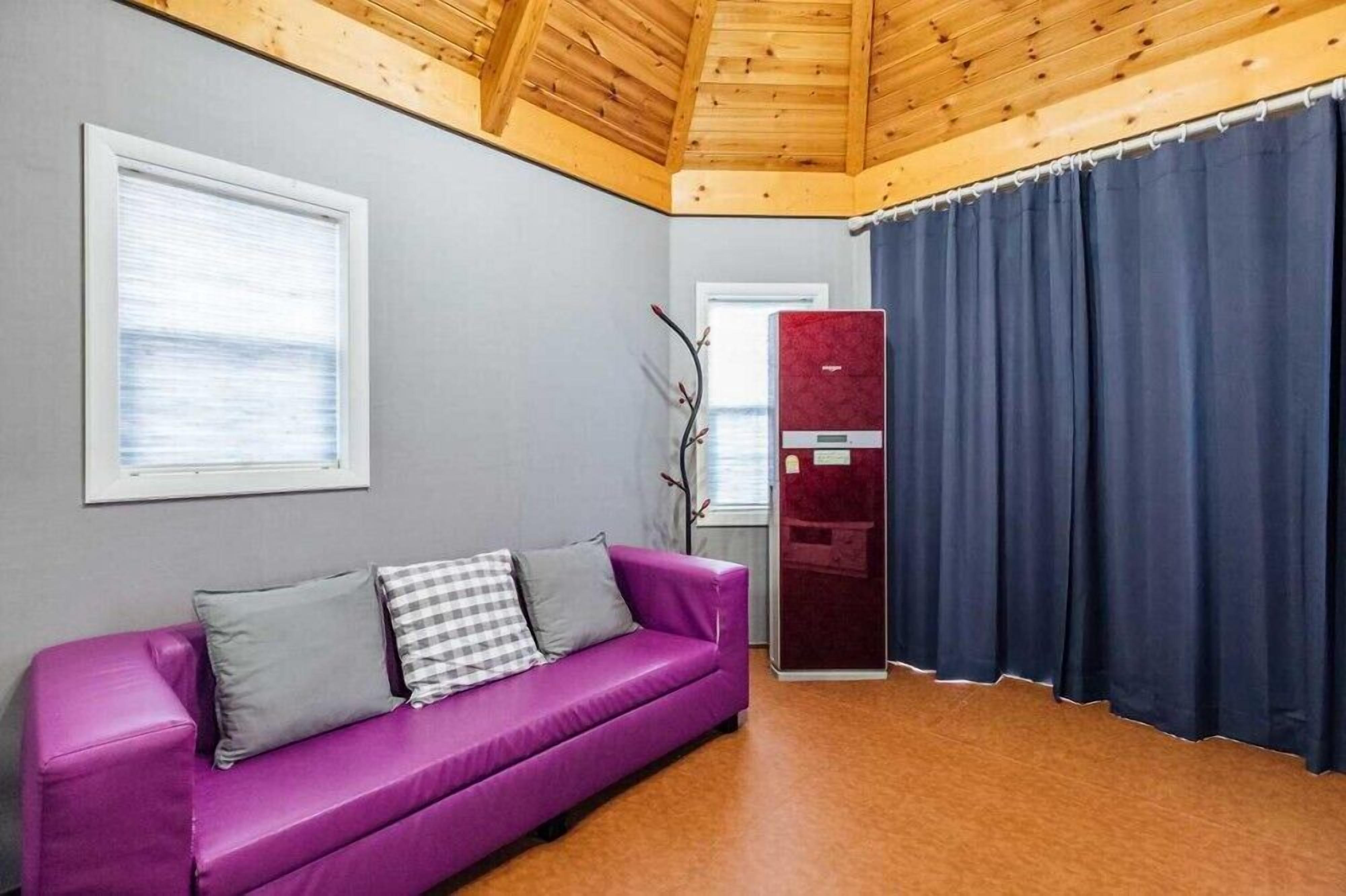 Una habitación con un sofá morado, una ventana con persianas, una cortina oscura y un techo de madera.