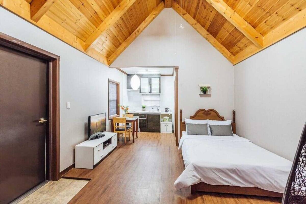 A bedroom with a wooden ceiling, a bed, a desk, and a kitchenette.