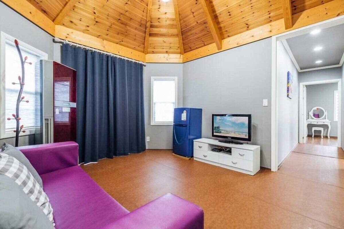 A room with a wooden ceiling, a television, a blue refrigerator, a purple sofa, and a white desk.