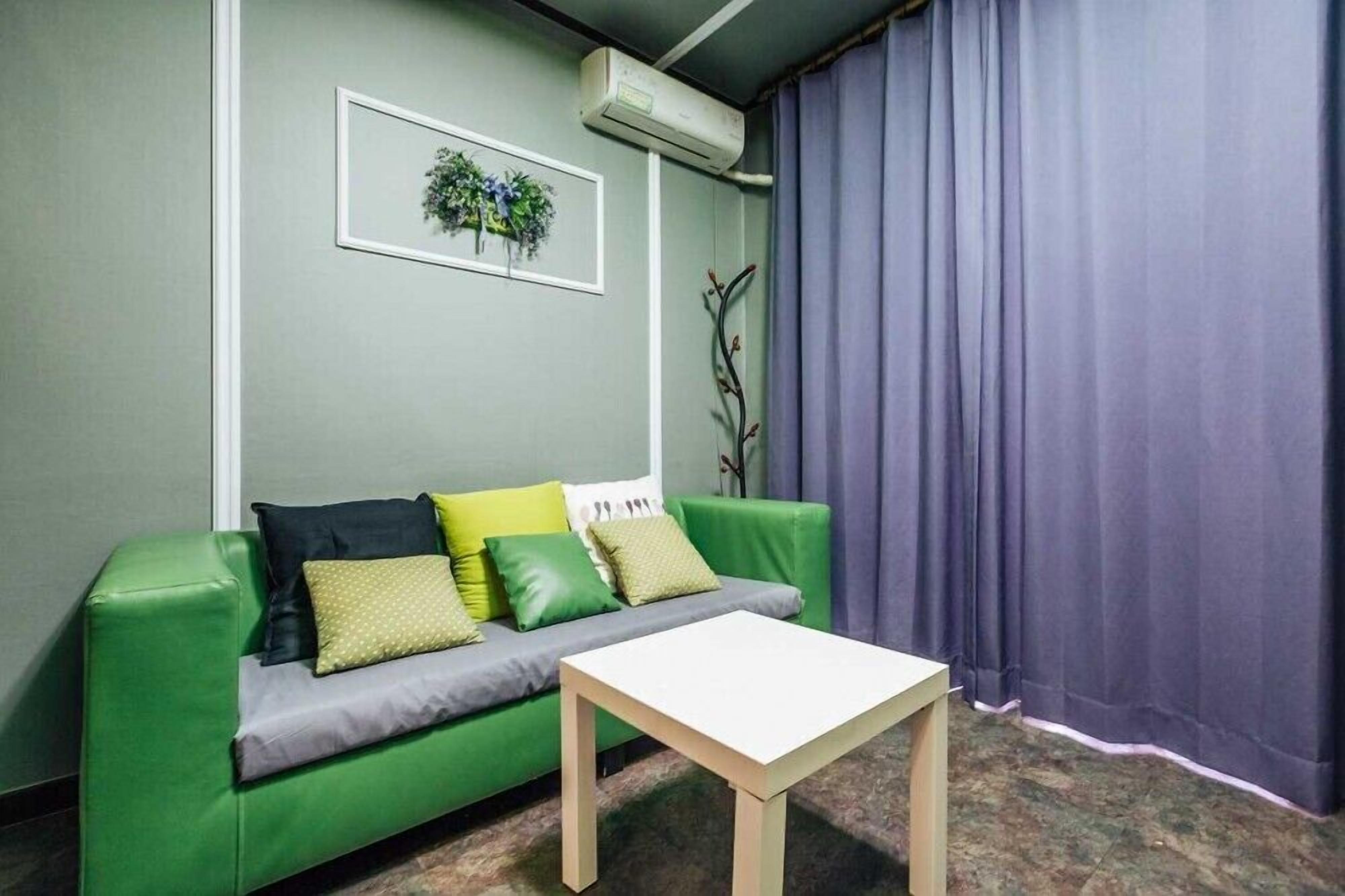 A room with a green sofa, a small white table, a wall-mounted air conditioner, and a window with purple curtains.