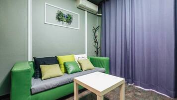 A room with a green sofa, a small white table, a wall-mounted air conditioner, and a window with purple curtains.