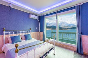 Une chambre avec un lit, une fenêtre donnant sur les montagnes et l’eau, et une balustrade de balcon.