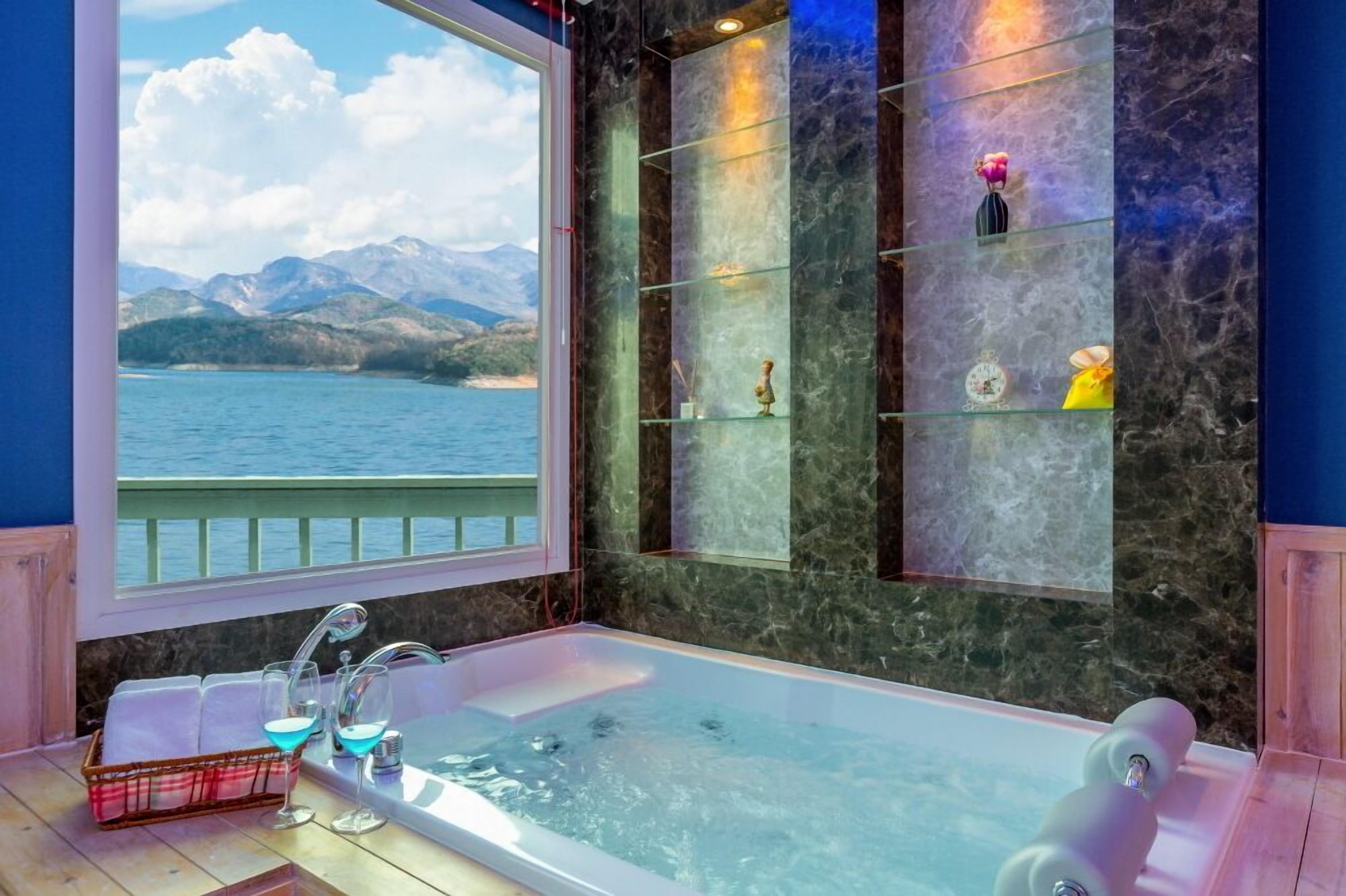 Une salle de bain avec une baignoire, une fenêtre donnant sur les montagnes et un lac, ainsi que des étagères ornées d’objets décoratifs.