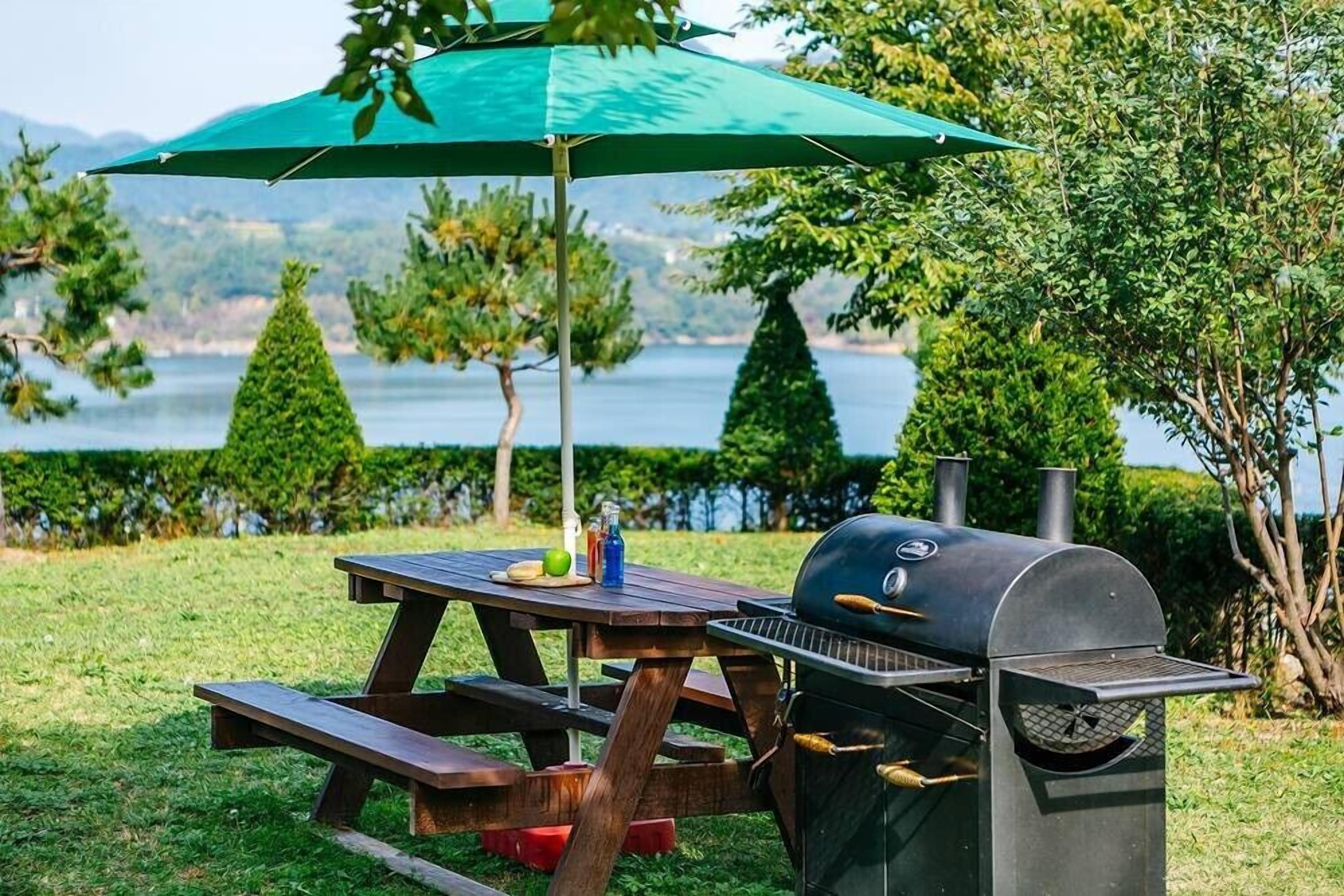 A picnic setup with a grill, table, and umbrella in a grassy area with trees and a lake in the background.