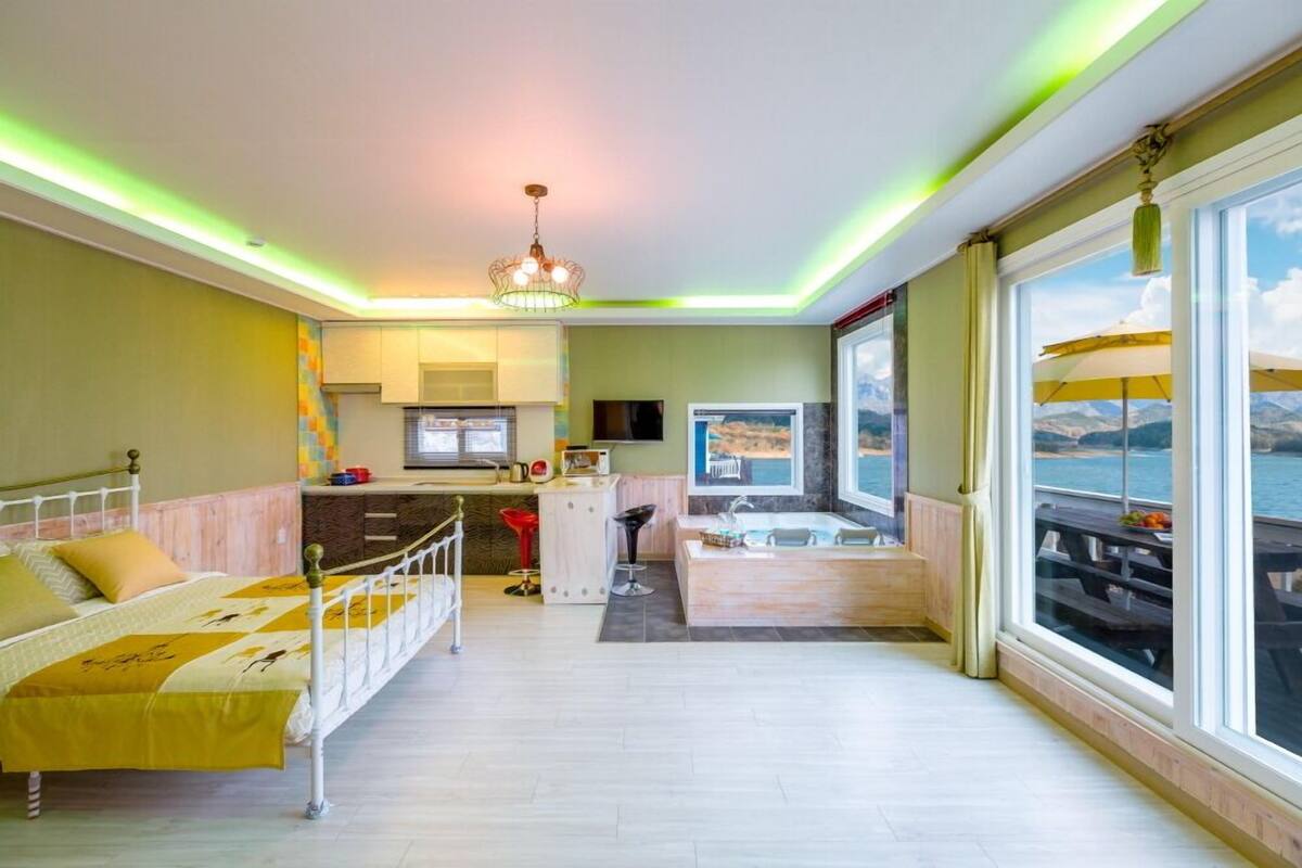 A room with a bed, a small kitchenette, a bathroom with a bathtub, and large windows with a view of the sea.