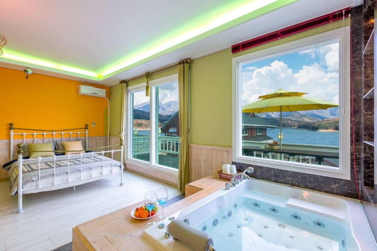 A room with a bed, a bathtub, and a large window with a view of mountains and water.
