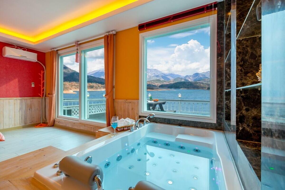 A room with a large bathtub, a window with a view of mountains and water, and a wooden floor.