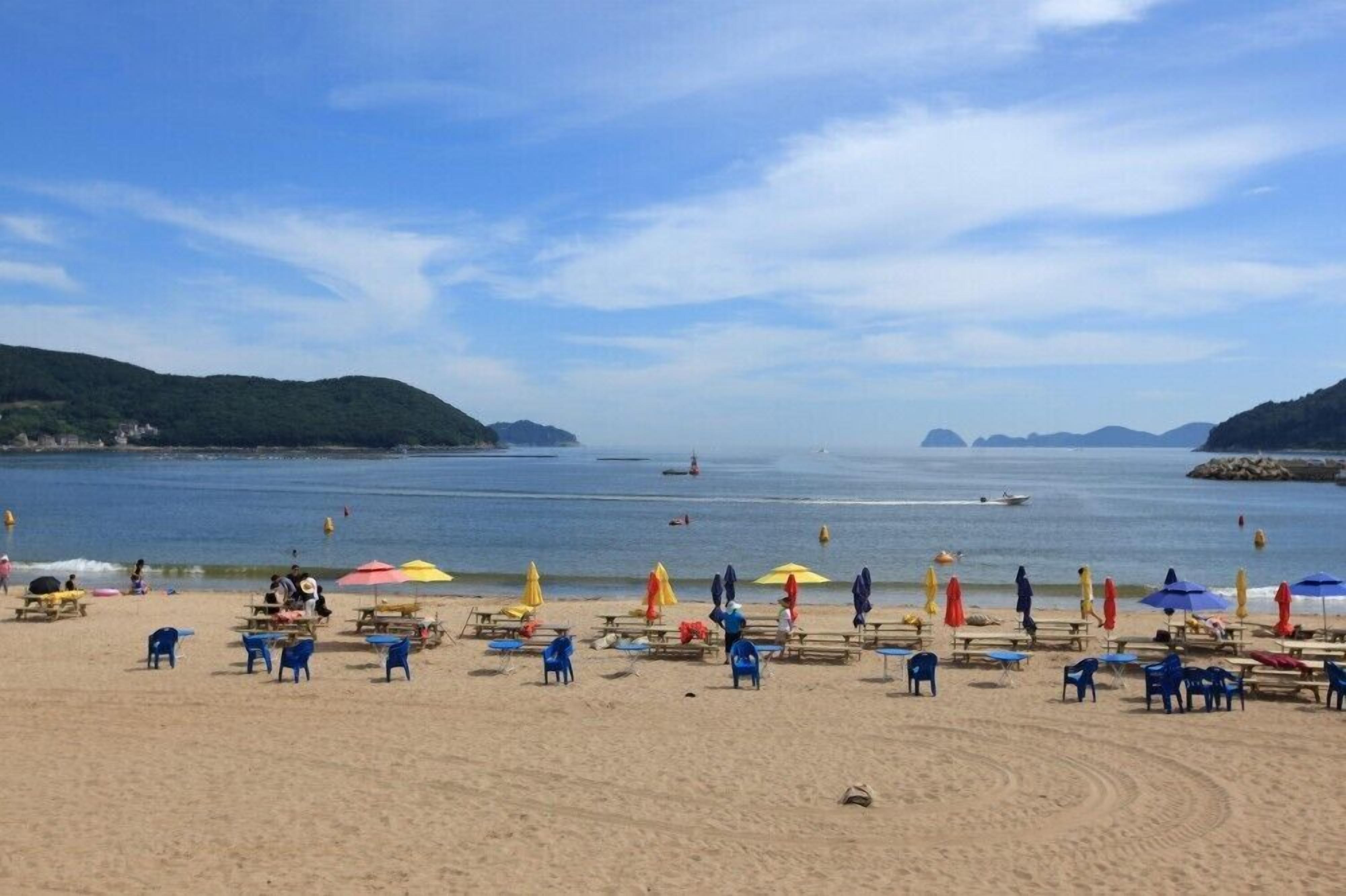 A beach with rows of blue chairs and umbrellas, a sandy shore, and calm water with boats.
