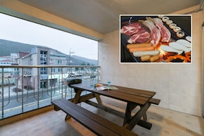 A balcony with a wooden table and benches, overlooking a street with buildings and a hilly landscape. - Gatnaeum Pension (Geoje)