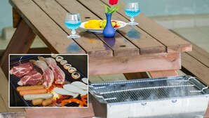 Una mesa de madera con un jarrón azul y dos vasos de líquido azul, un plato de fruta, y una parrilla con carne y salchichas.