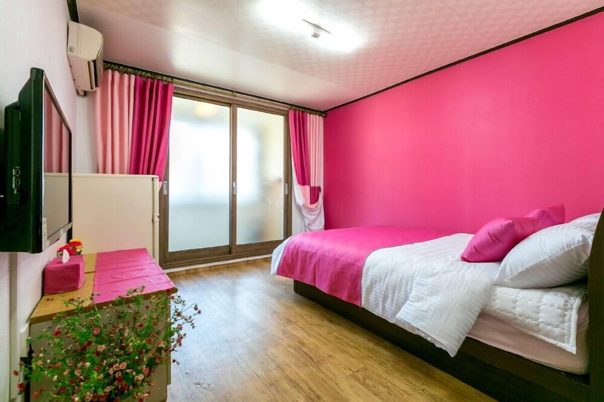 A bedroom with a pink wall, a bed with white bedding, a wooden floor, a vanity with a mirror, and a window with pink curtains.
