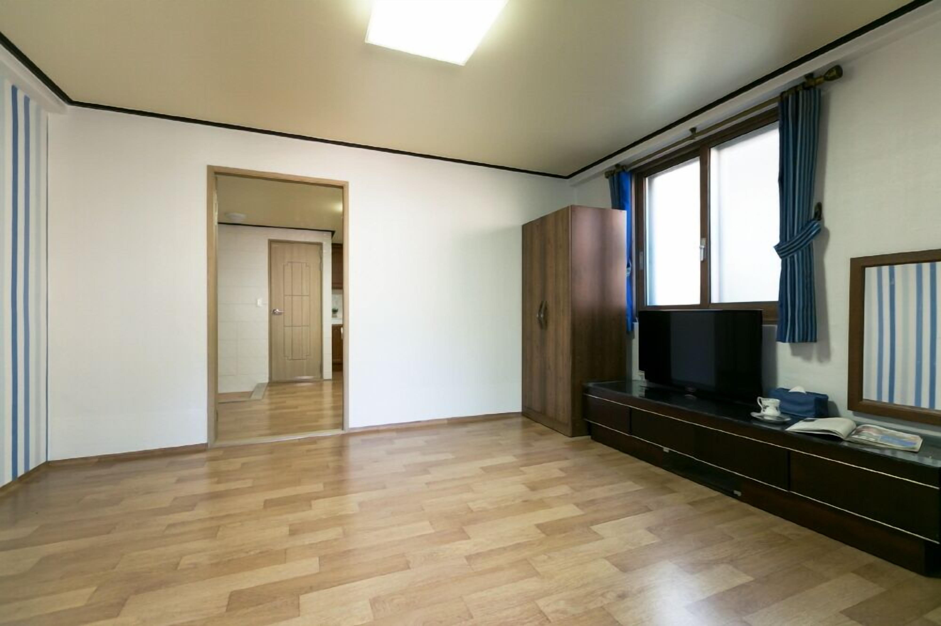 Quarto com piso de madeira, televisão sobre suporte, guarda-roupa e janela com cortinas.