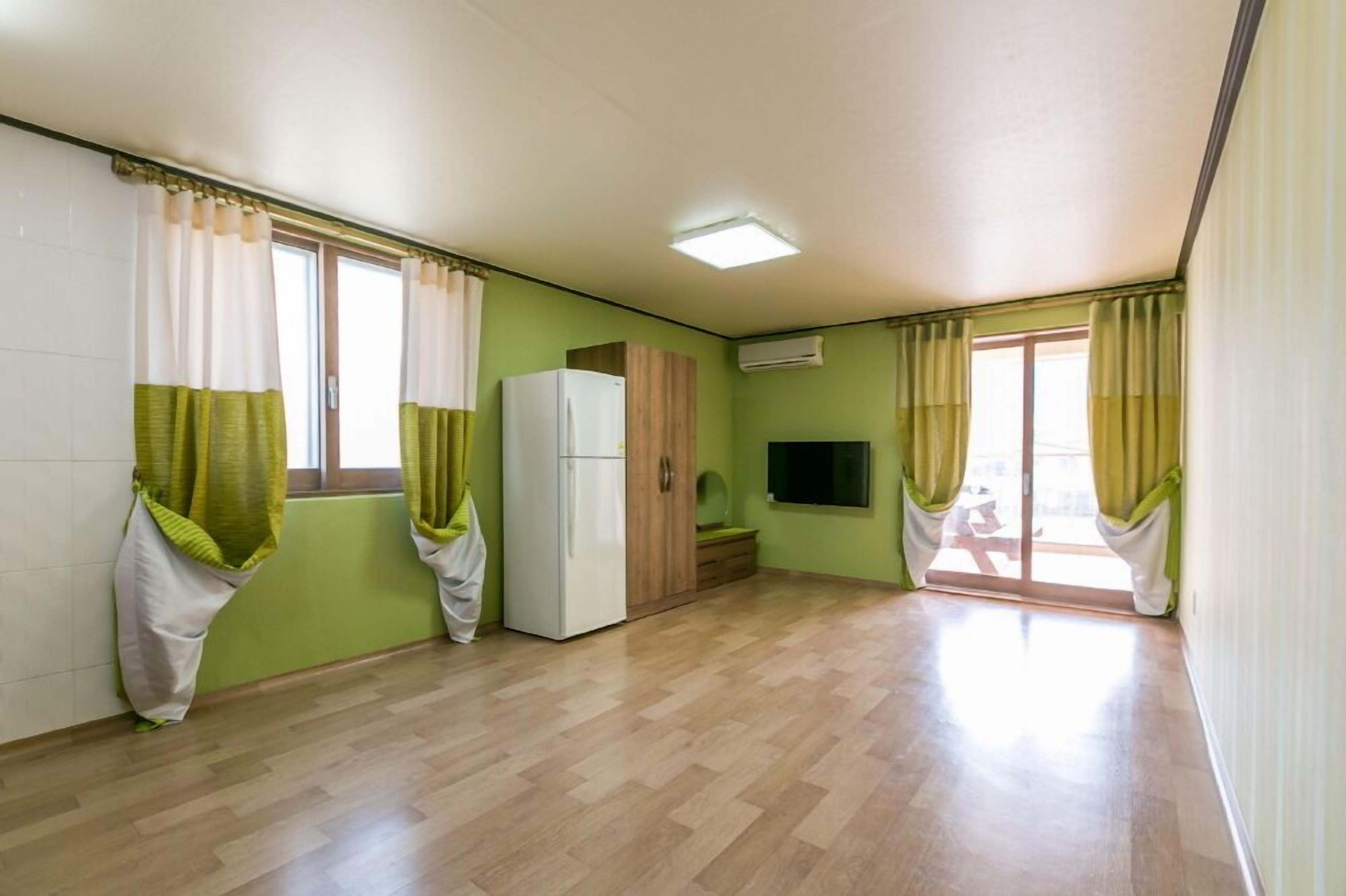 A room with a wooden floor, a white refrigerator, a wooden cabinet, a flat-screen TV, and a sliding glass door leading to a balcony.