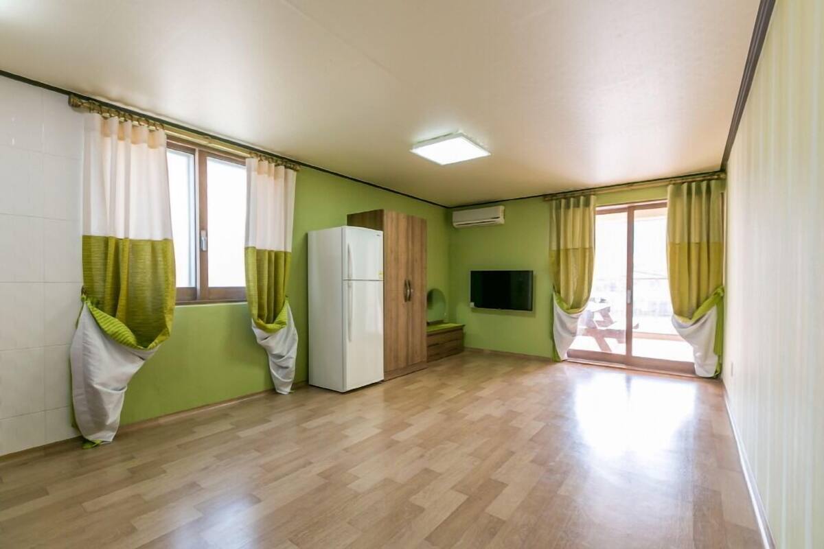 A room with a wooden floor, a white refrigerator, a wooden cabinet, a flat-screen TV, and a sliding glass door leading to a balcony.