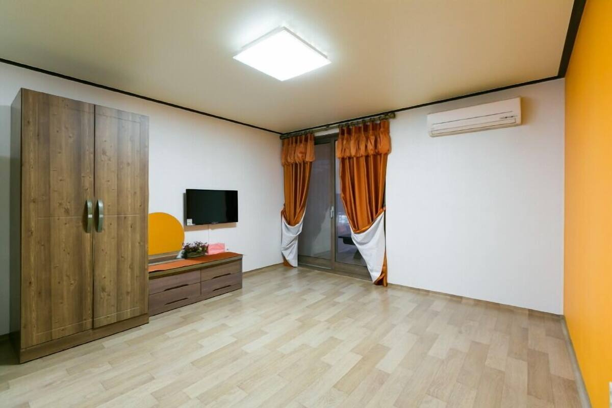 A room with a wardrobe, a flat-screen TV, a desk with a pink object, and orange curtains.