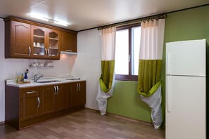 A compact kitchen with wooden cabinets, a white refrigerator, and a window with green curtains. - Gatnaeum Pension (Geoje)