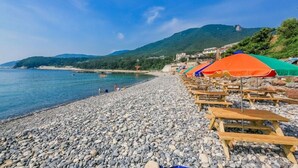 A pebble beach with wooden benches and colorful umbrellas, overlooking a calm sea and green hills. - Gaejul Trip Pension (Geoje)
