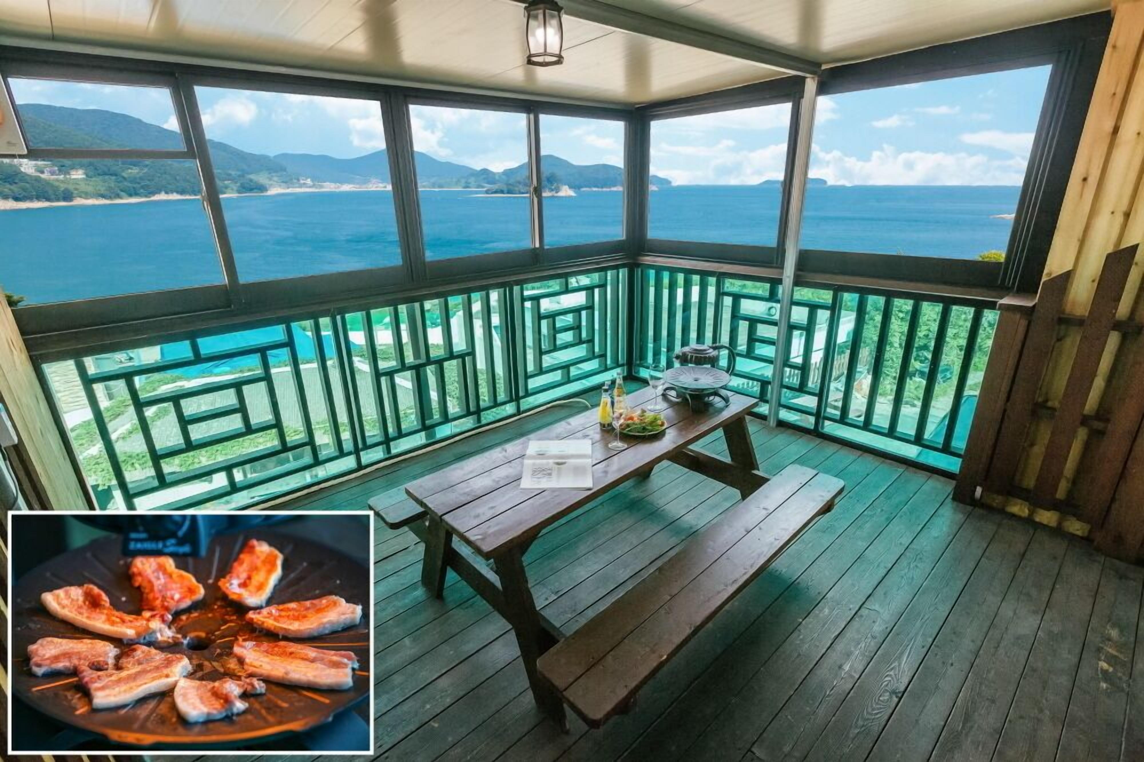 A covered outdoor area with a wooden table and benches, overlooking a coastal view with mountains and the sea.