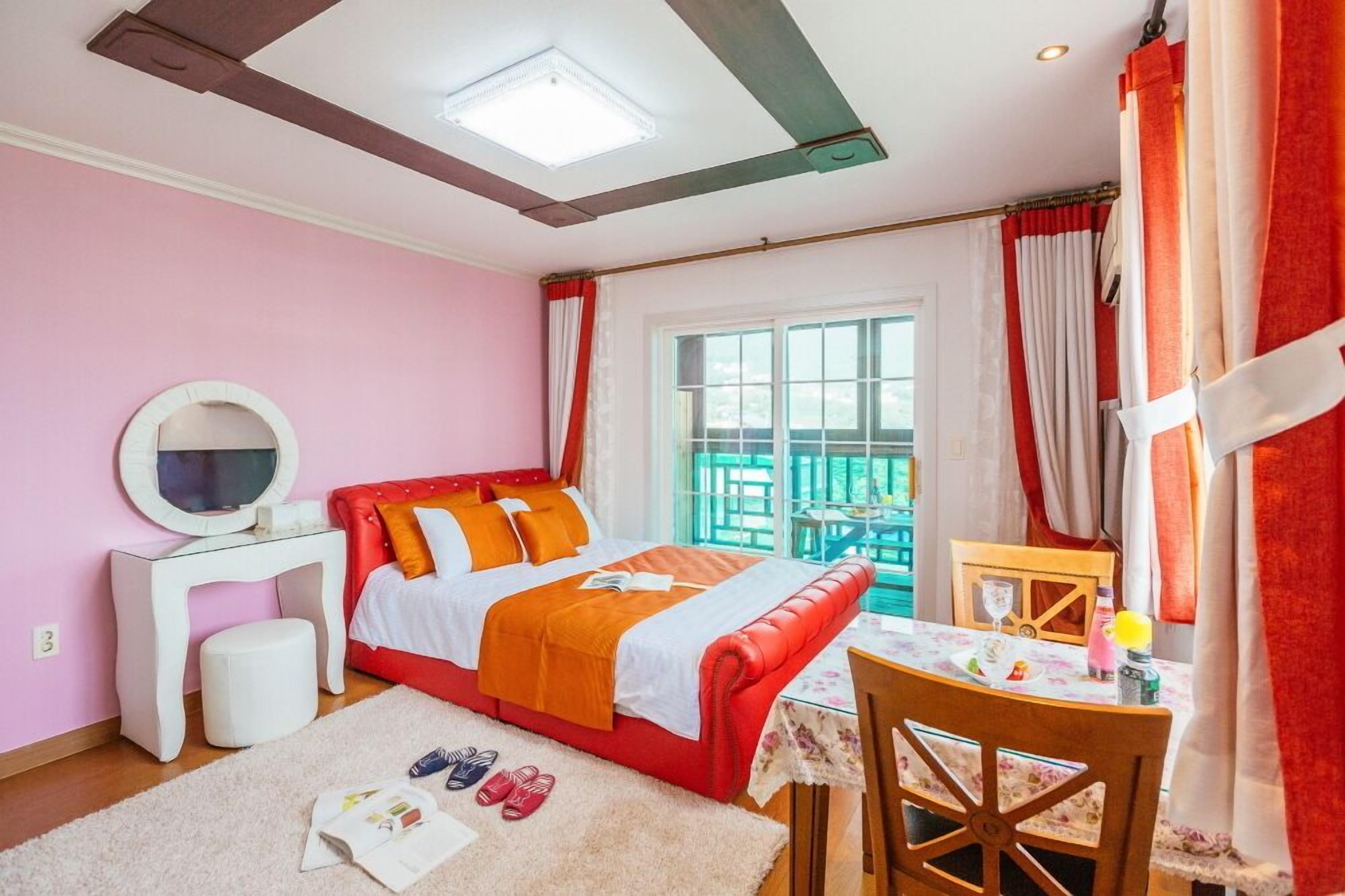 A bedroom with a bed, a bedside table, a mirror, a window with curtains, and a small table with a chair.