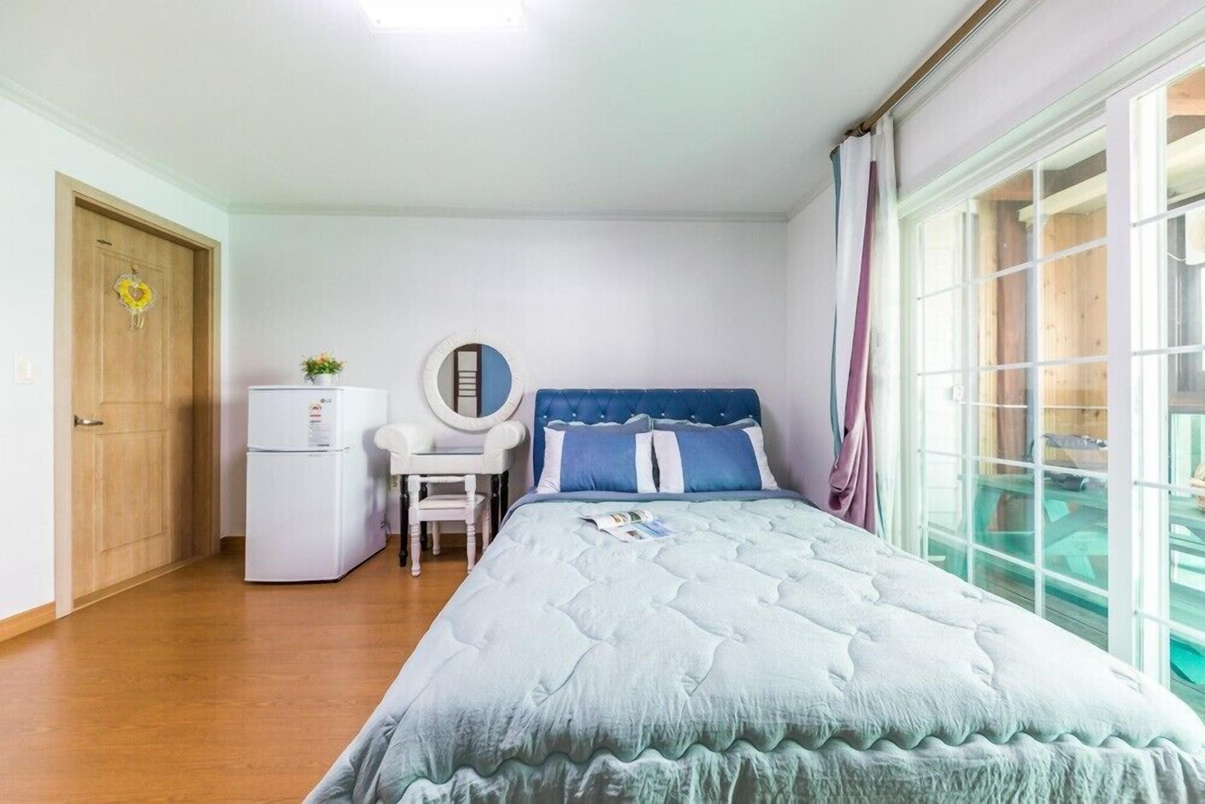 A bedroom with a bed, a desk, a chair, a small refrigerator, and a window with curtains.
