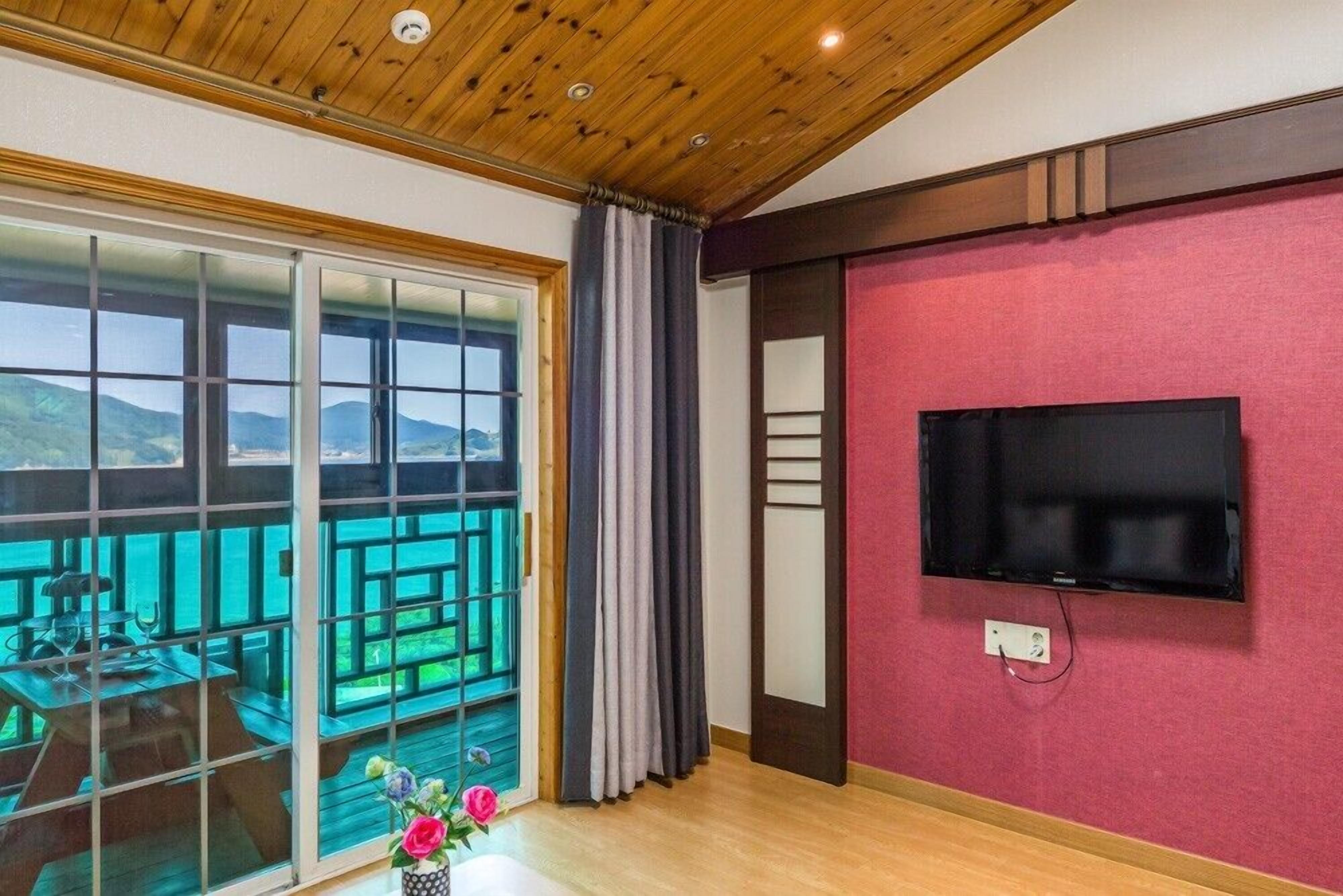A room with a wooden ceiling, a TV mounted on a red wall, a sliding glass door leading to a balcony with a view of mountains, and a wooden table with a vase of flowers.