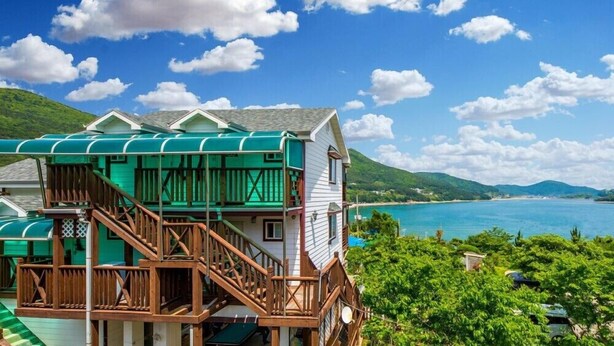 A two-story building with a green roof and wooden balcony, overlooking a turquoise sea and green hills. - Gaejul Trip Pension (Geoje)
