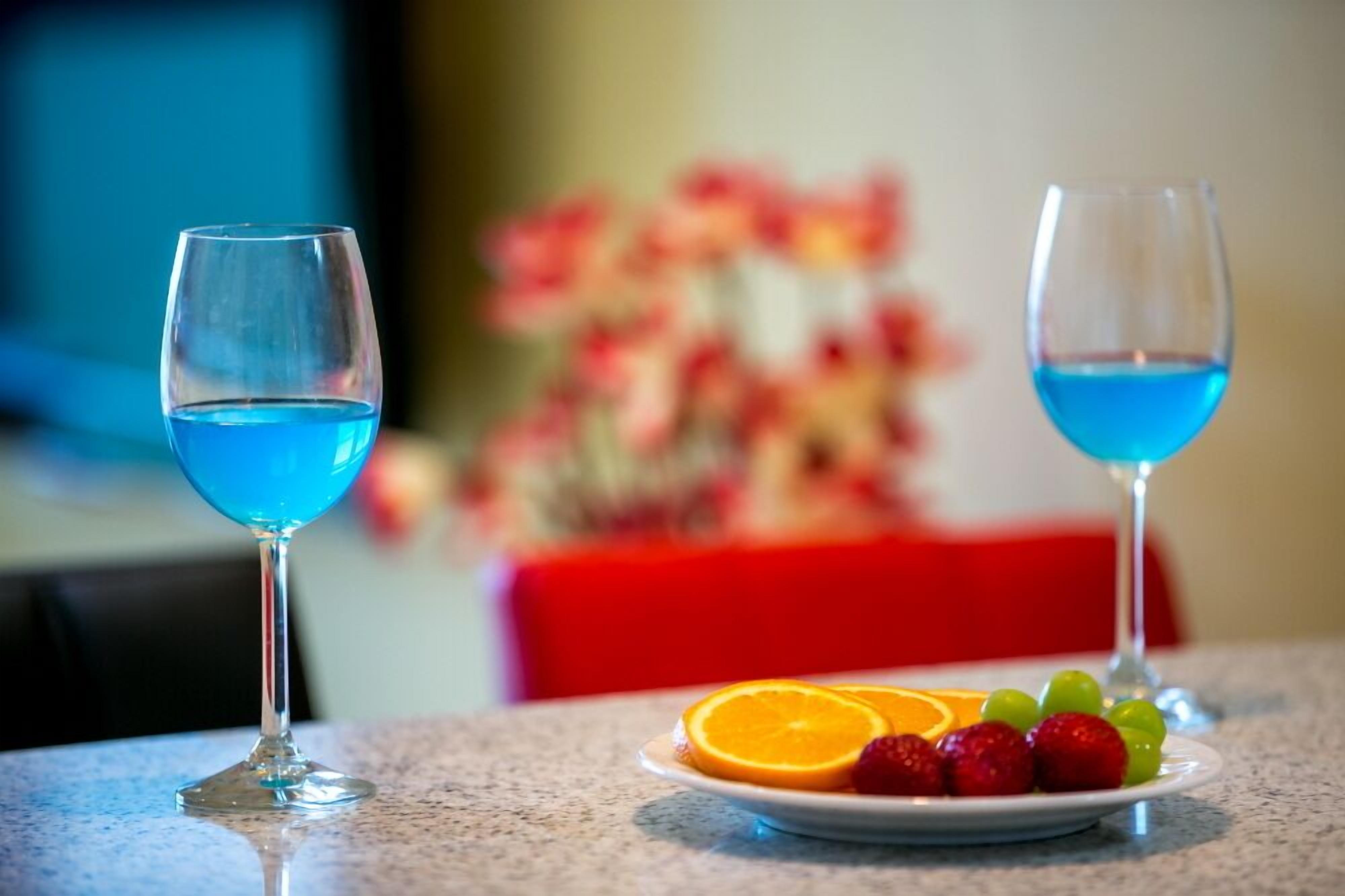 Deux verres contenant un liquide bleu sont posés sur une table, avec une assiette de fruits en premier plan.