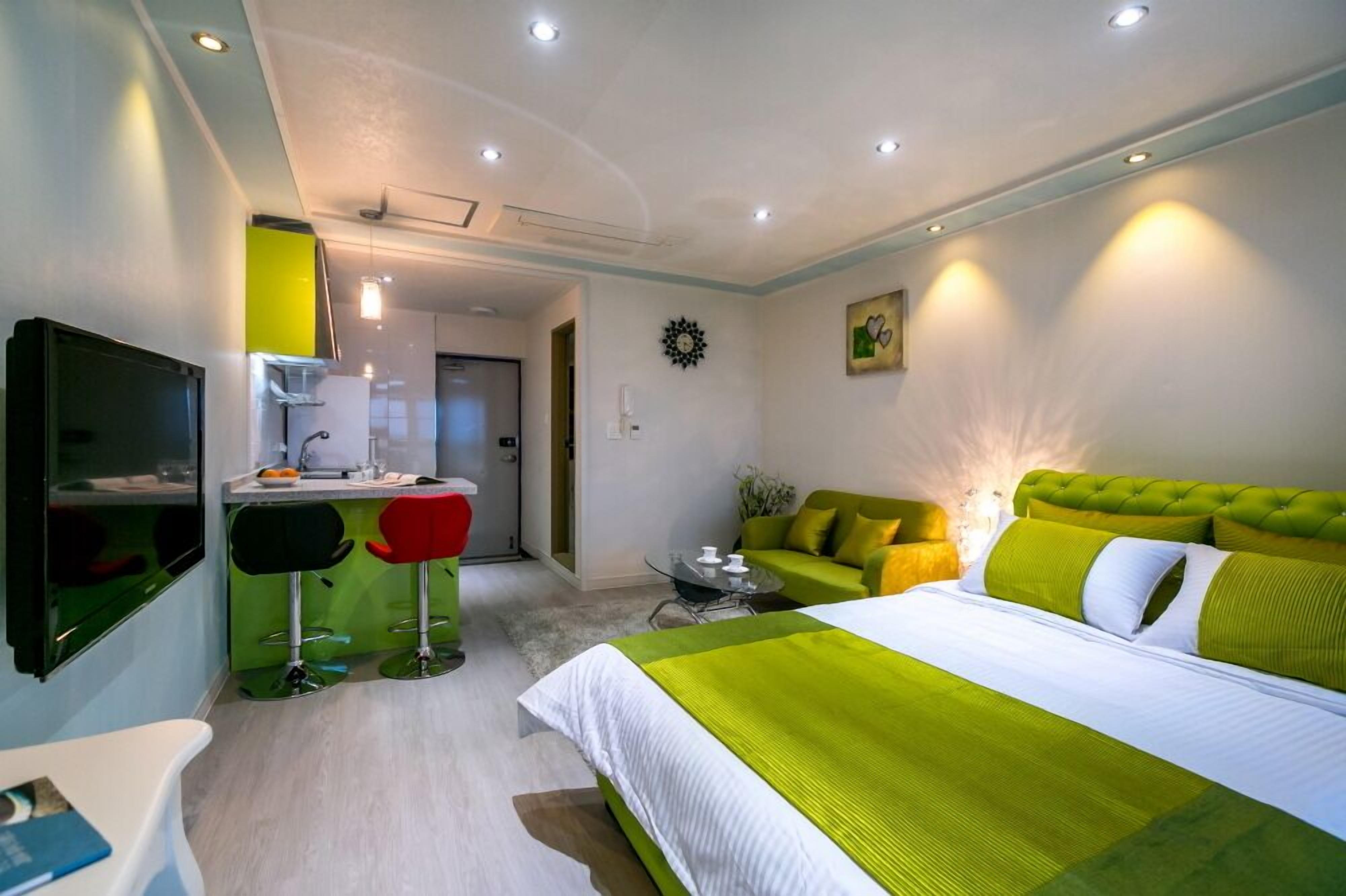 A modern hotel room with a large bed, a desk with a chair, a TV, and a dining area with stools.