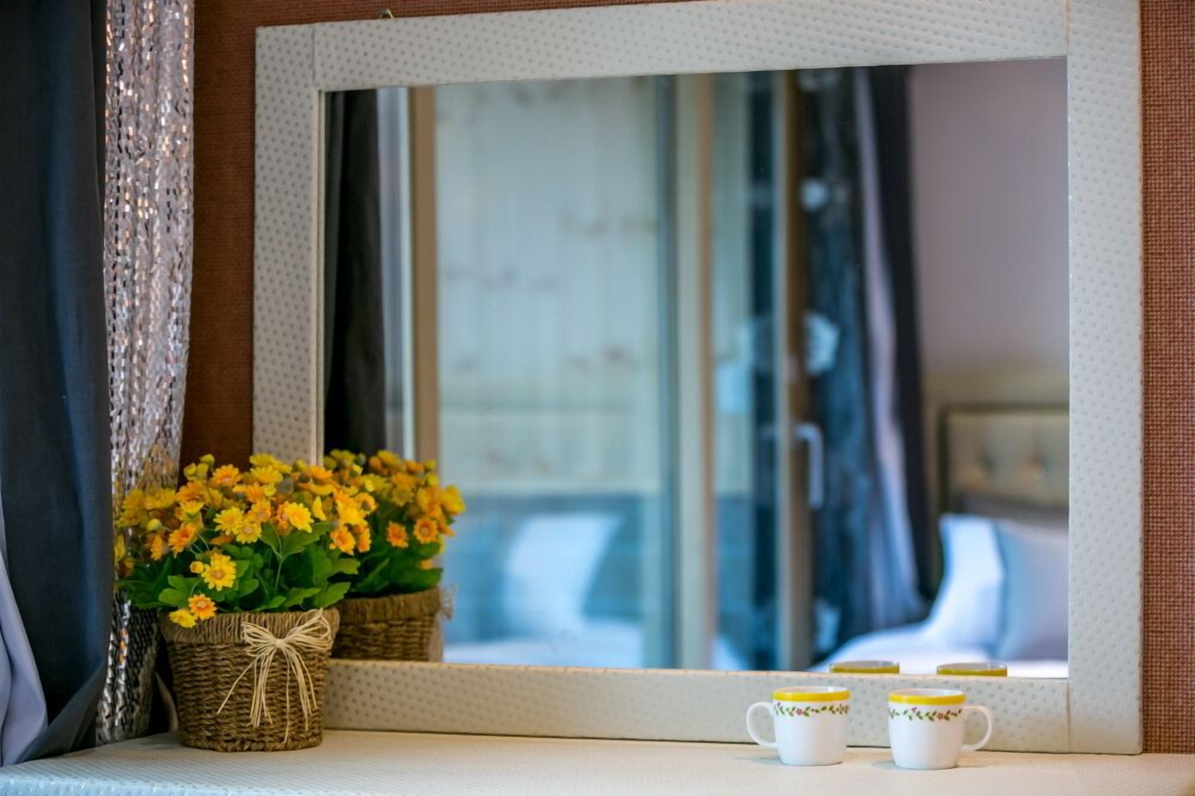 A mirror with a reflection of a window, a small basket of yellow flowers, and three white mugs with yellow lids on a table.