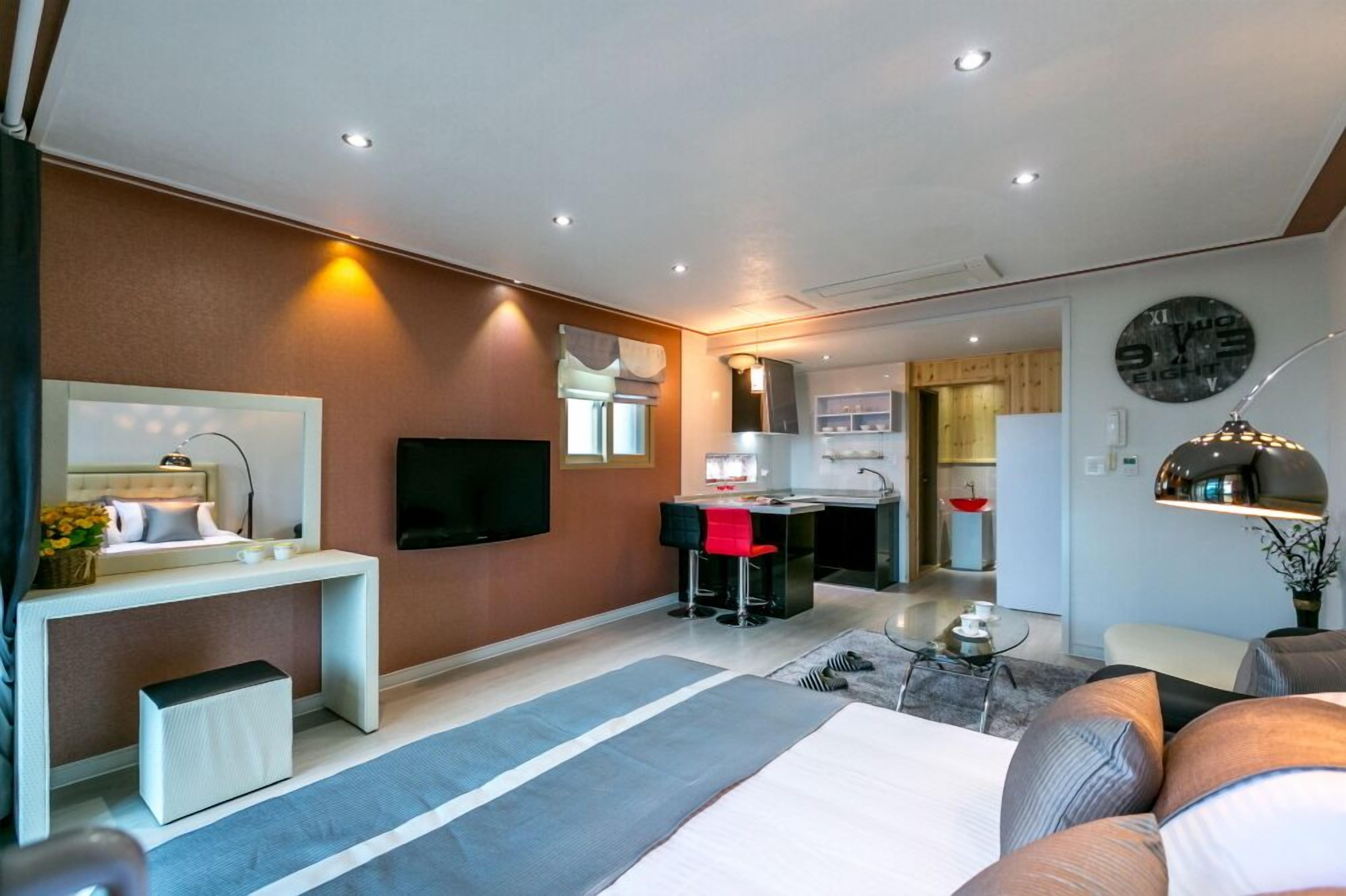 A modern hotel room with a bed, a desk, a TV, and a kitchen area in the background.