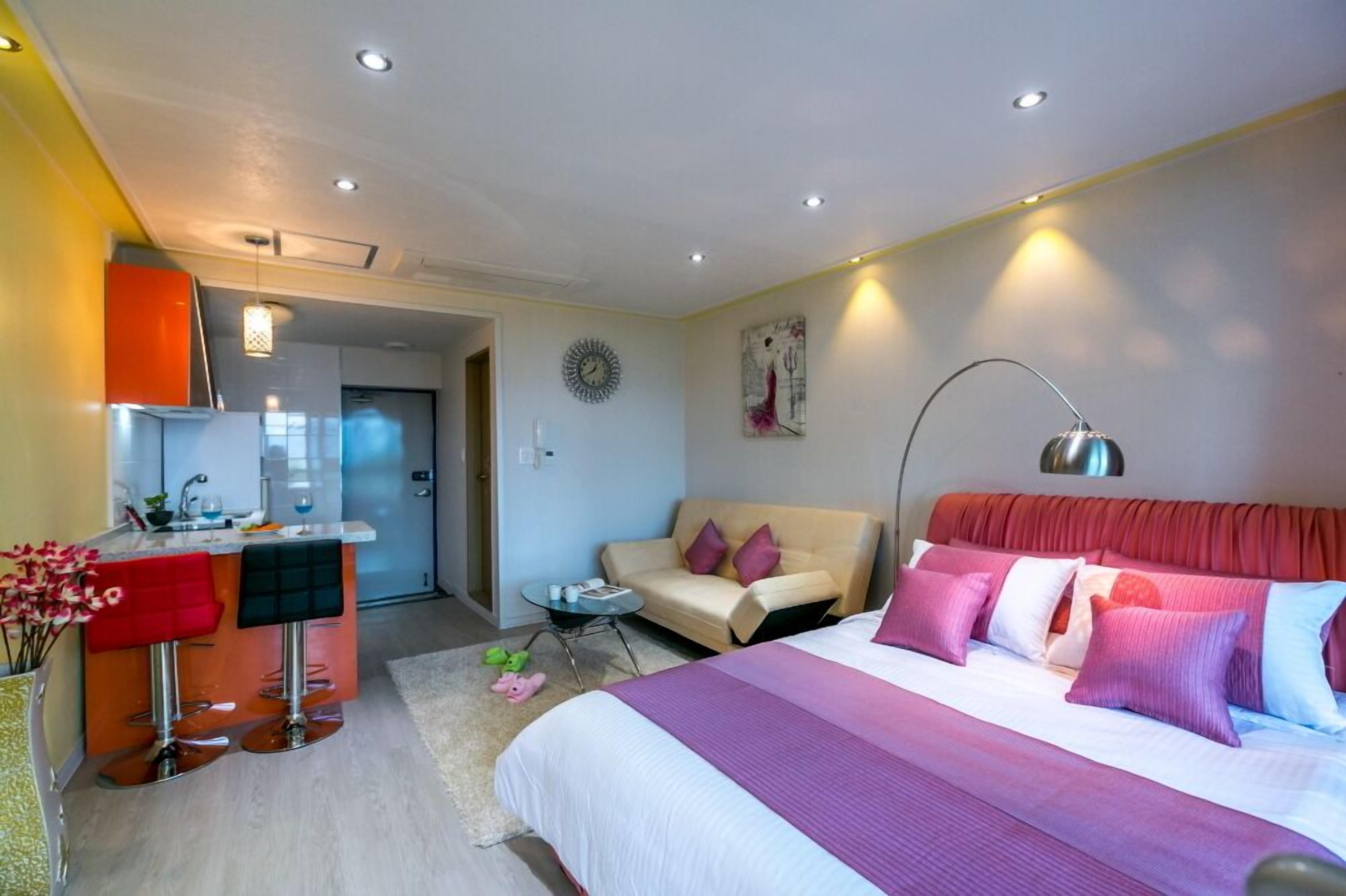 A modern hotel room with a bed, a sofa, a small table, and a kitchenette.