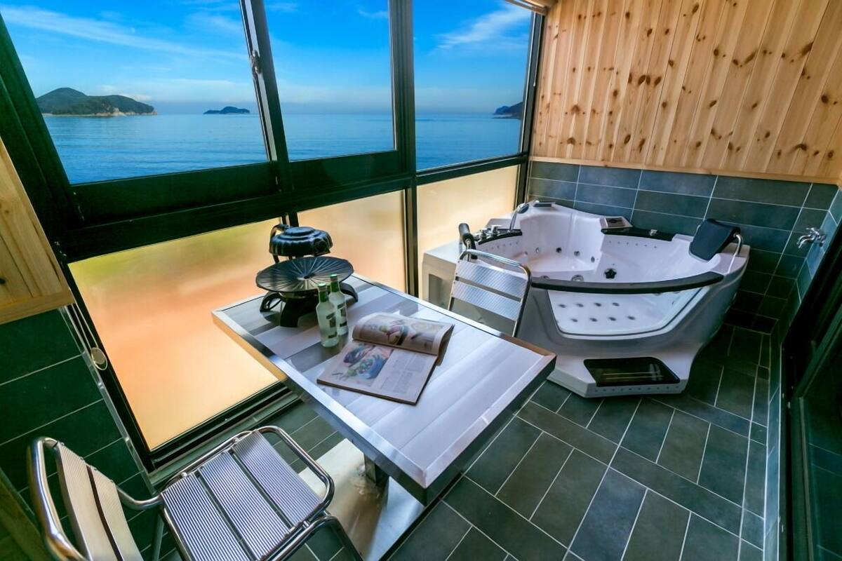 Una habitación con un ventanal grande que ofrece vista al mar, una bañera, una mesa con una revista y una pequeña zona de cocina.