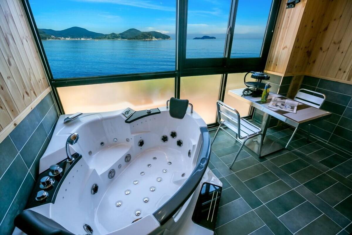 Un baño moderno con una bañera grande, una mesita con una silla y vistas al mar a través de amplios ventanales.