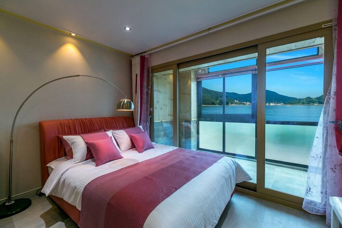 Un dormitorio con una cama grande, una lámpara de pie y vistas a un cuerpo de agua a través de una puerta corrediza.