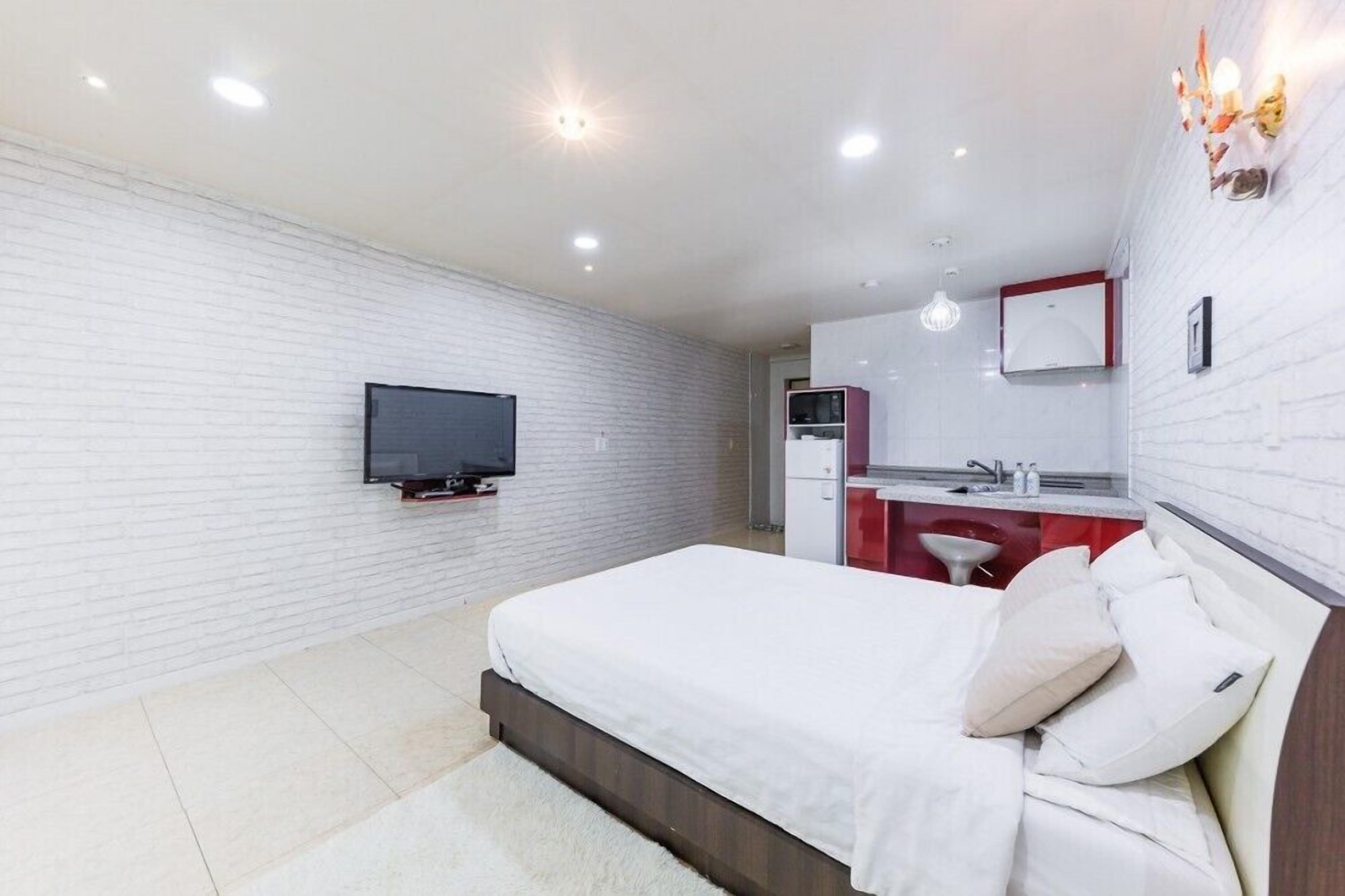 A modern bedroom with a bed, a TV mounted on the wall, a red and white kitchenette, and a white brick wall.