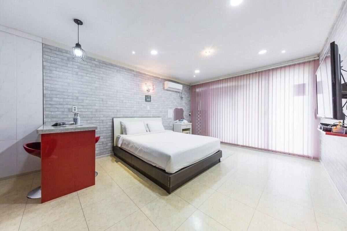 A modern bedroom with a bed, a red cabinet, a white bedside table, and a large window with blinds.